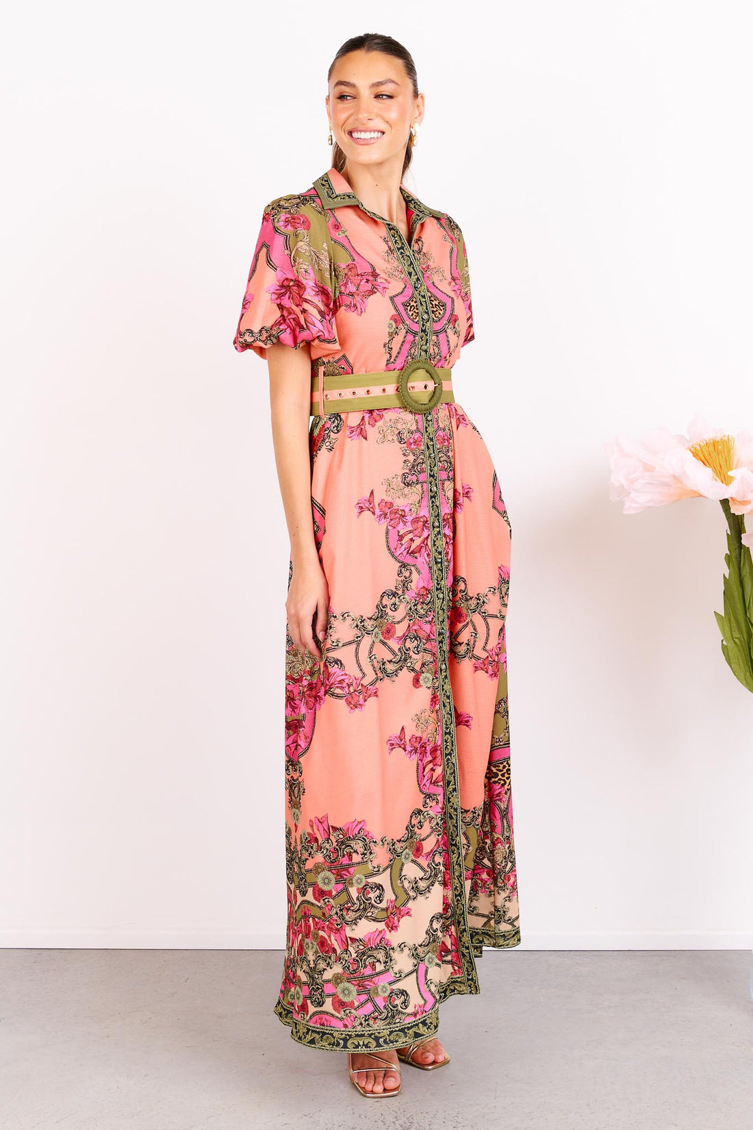 Woman wearing the minnie + MAXXIE Madison Garden peachy pink floral maxi event dress with a crochet buckle belt, standing against a white background.