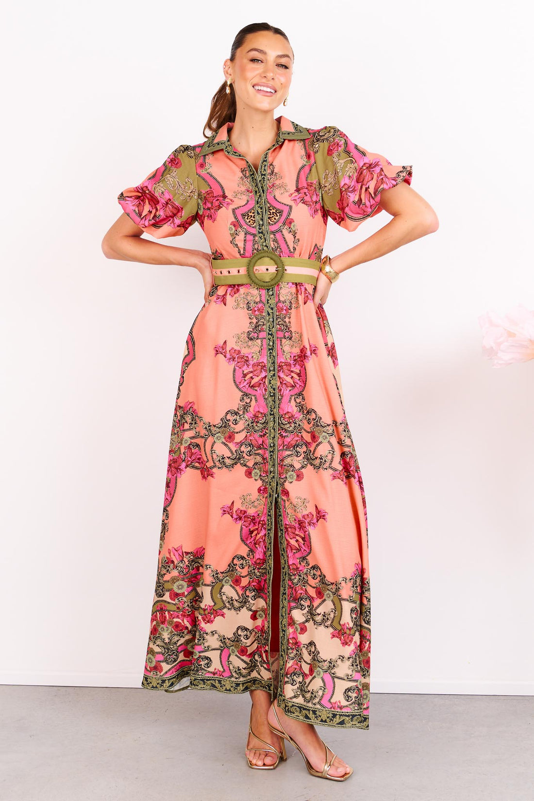 Woman wearing the minnie + MAXXIE Madison Garden peachy pink floral maxi event dress with a crochet buckle belt, standing against a white background.
