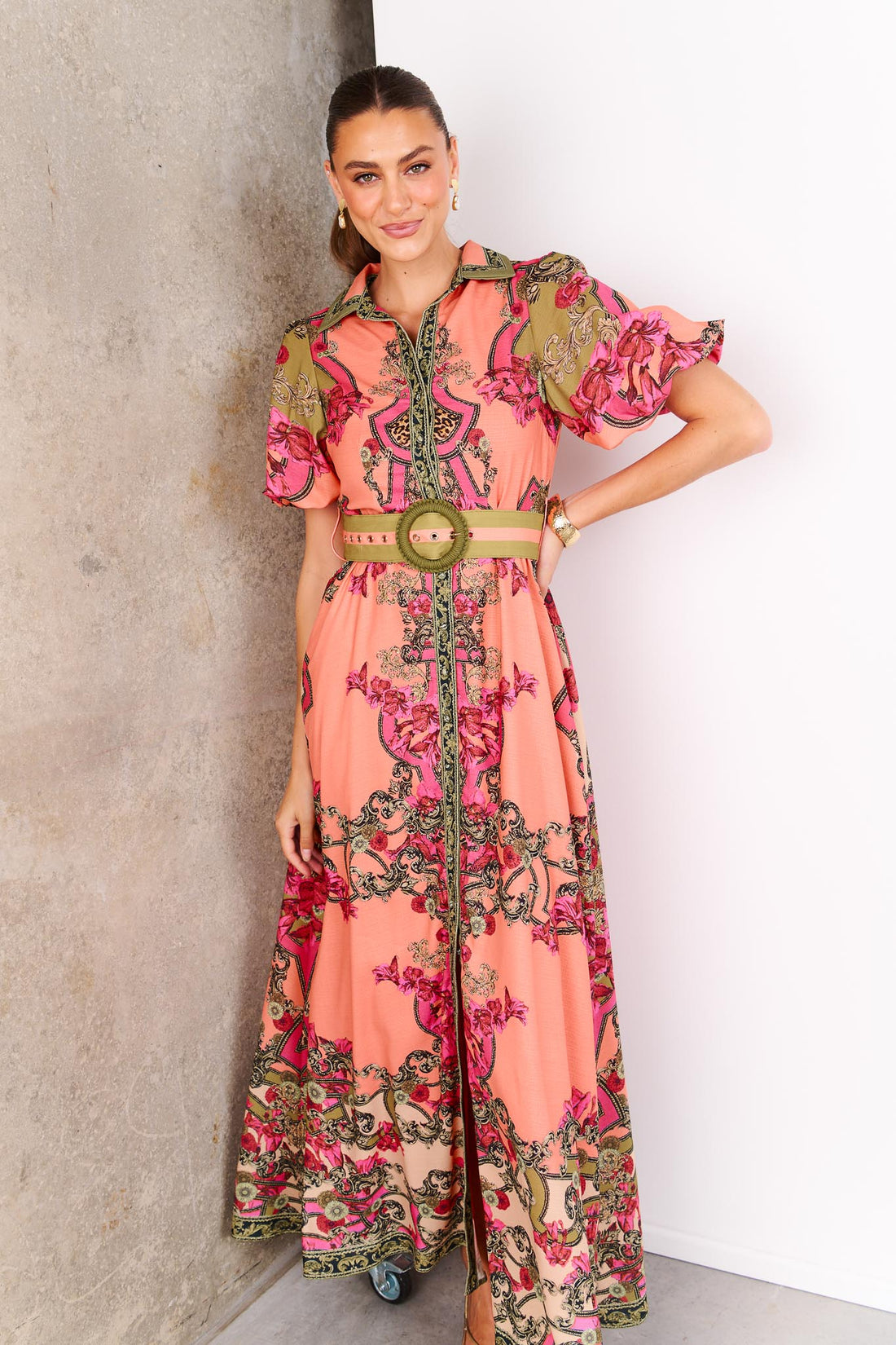 Woman wearing the minnie + MAXXIE Madison Garden peachy pink floral maxi event dress with a crochet buckle belt, standing against a white and concrete background.