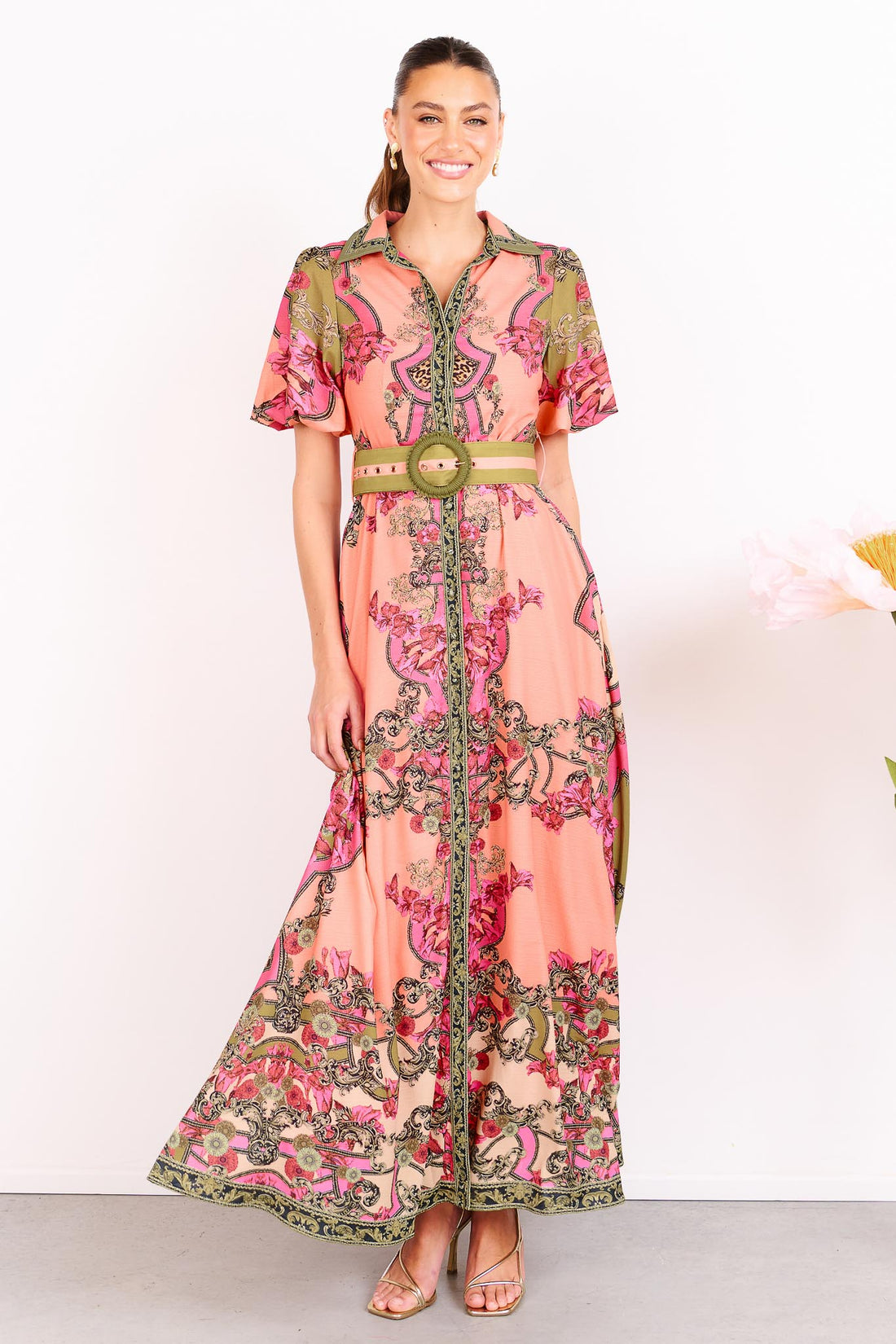 Woman wearing the minnie + MAXXIE Madison Garden peachy pink floral maxi event dress with a crochet buckle belt, standing against a white background.