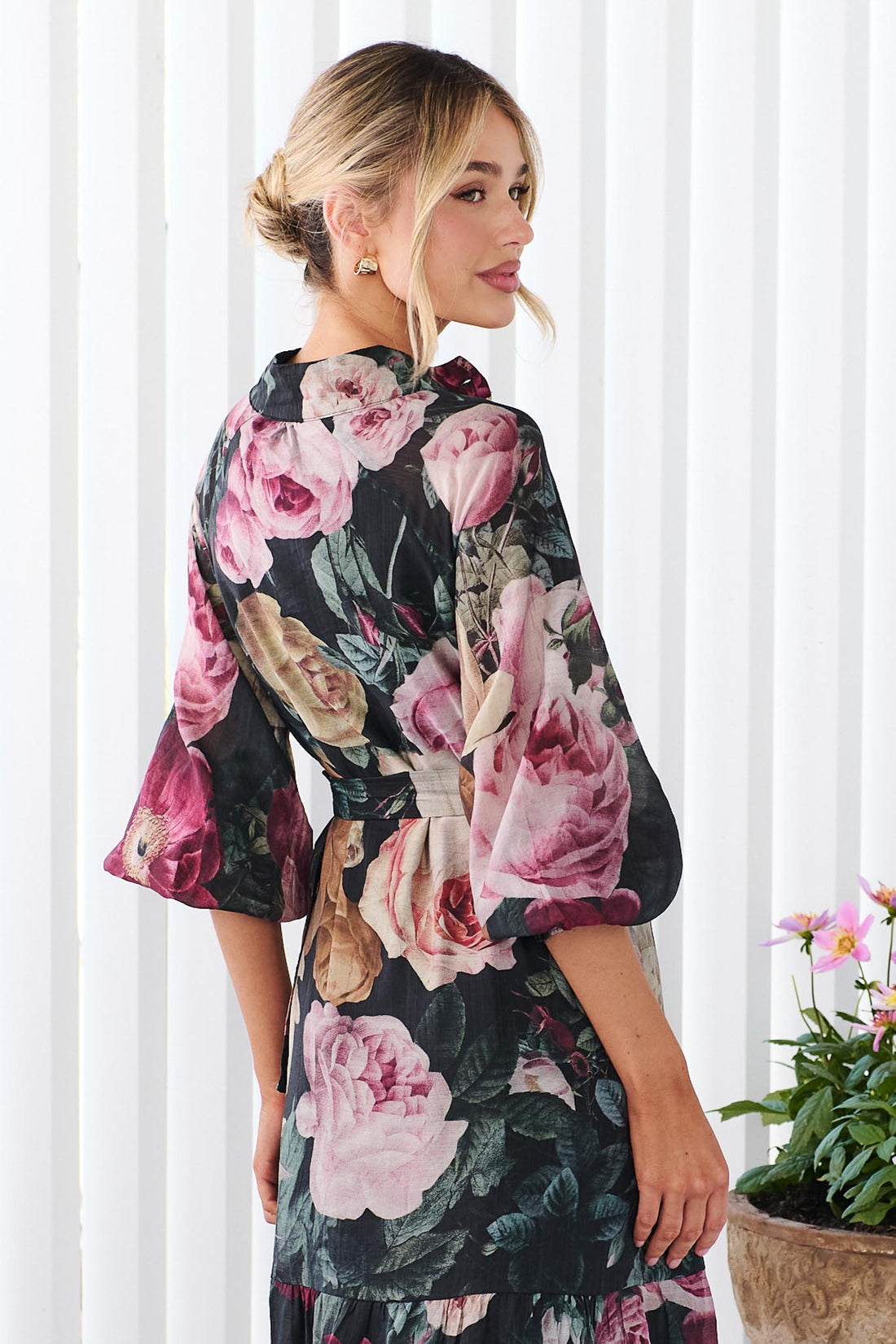A woman wearing the minnie + MAXXIE Maison long-sleeved maxi event dress with a dark floral print featuring pink, red, and beige flowers on a black background, standing in front of a white panelled wall.