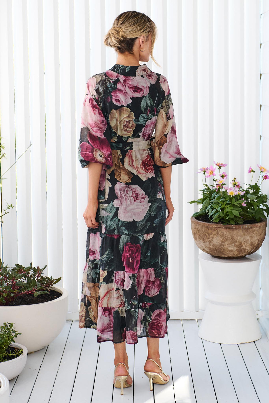Back view of a woman wearing the minnie + MAXXIE Maison long-sleeved maxi event dress with a dark floral print featuring pink, red, and beige flowers on a black background, standing in front of a white panelled wall.