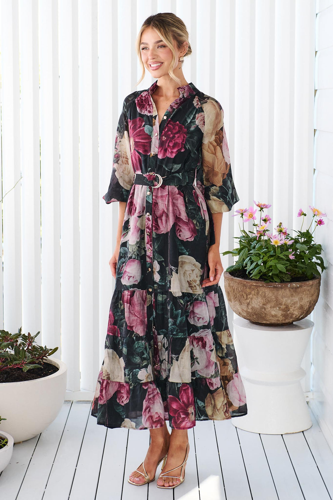 A woman wearing the minnie + MAXXIE Maison long-sleeved maxi event dress with a dark floral print featuring pink, red, and beige flowers on a black background, standing in front of a white panelled wall.