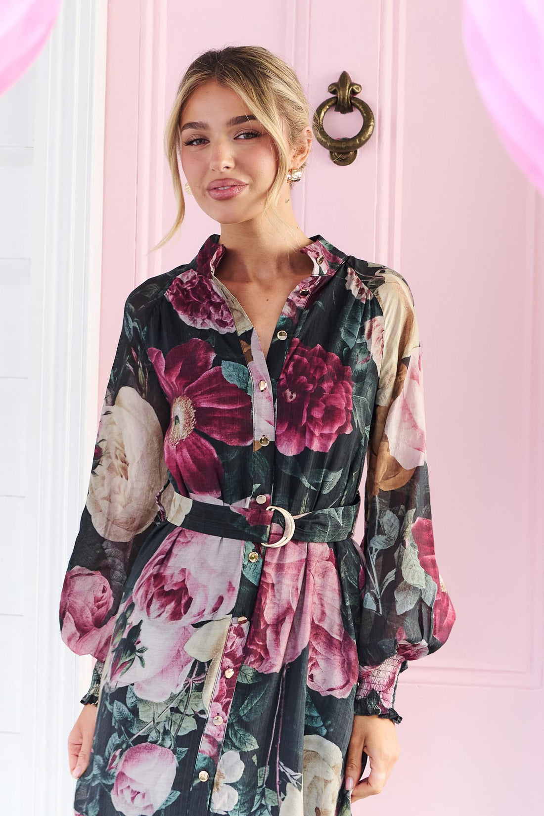 A woman wearing the minnie + MAXXIE Maison long-sleeved maxi event dress with a dark floral print featuring pink, red, and beige flowers on a black background, standing in front of a pink door.