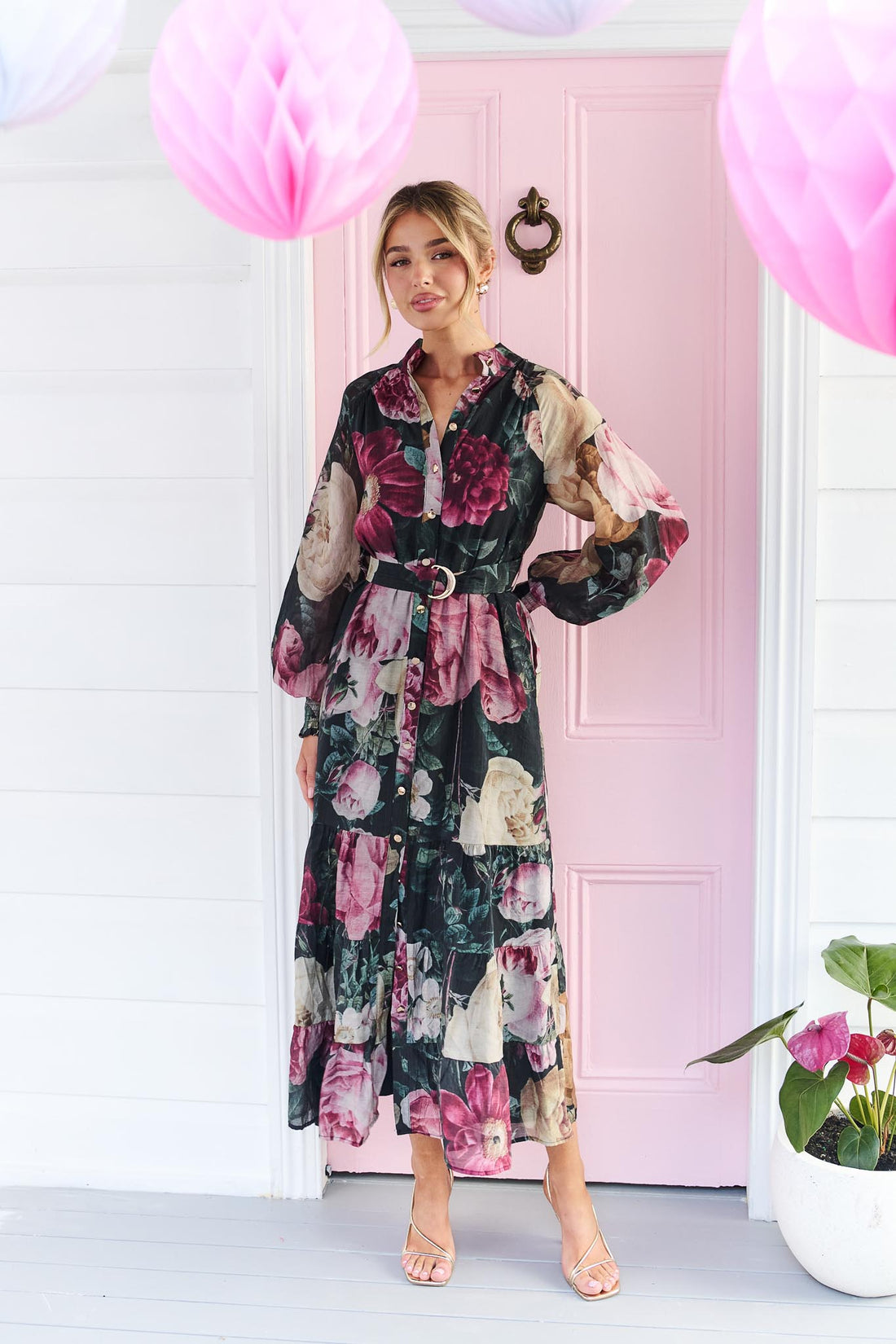 A woman wearing the minnie + MAXXIE Maison long-sleeved maxi event dress with a dark floral print featuring pink, red, and beige flowers on a black background, standing in front of a pink door.
