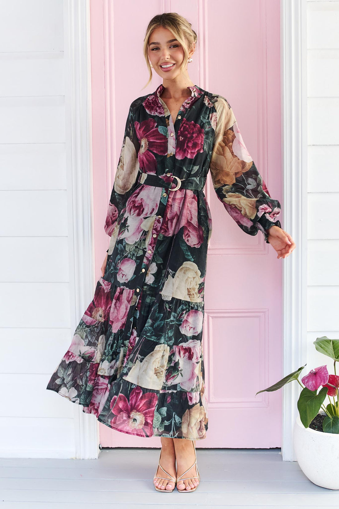 A woman wearing the minnie + MAXXIE Maison long-sleeved maxi event dress with a dark floral print featuring pink, red, and beige flowers on a black background, standing in front of a pink door.
