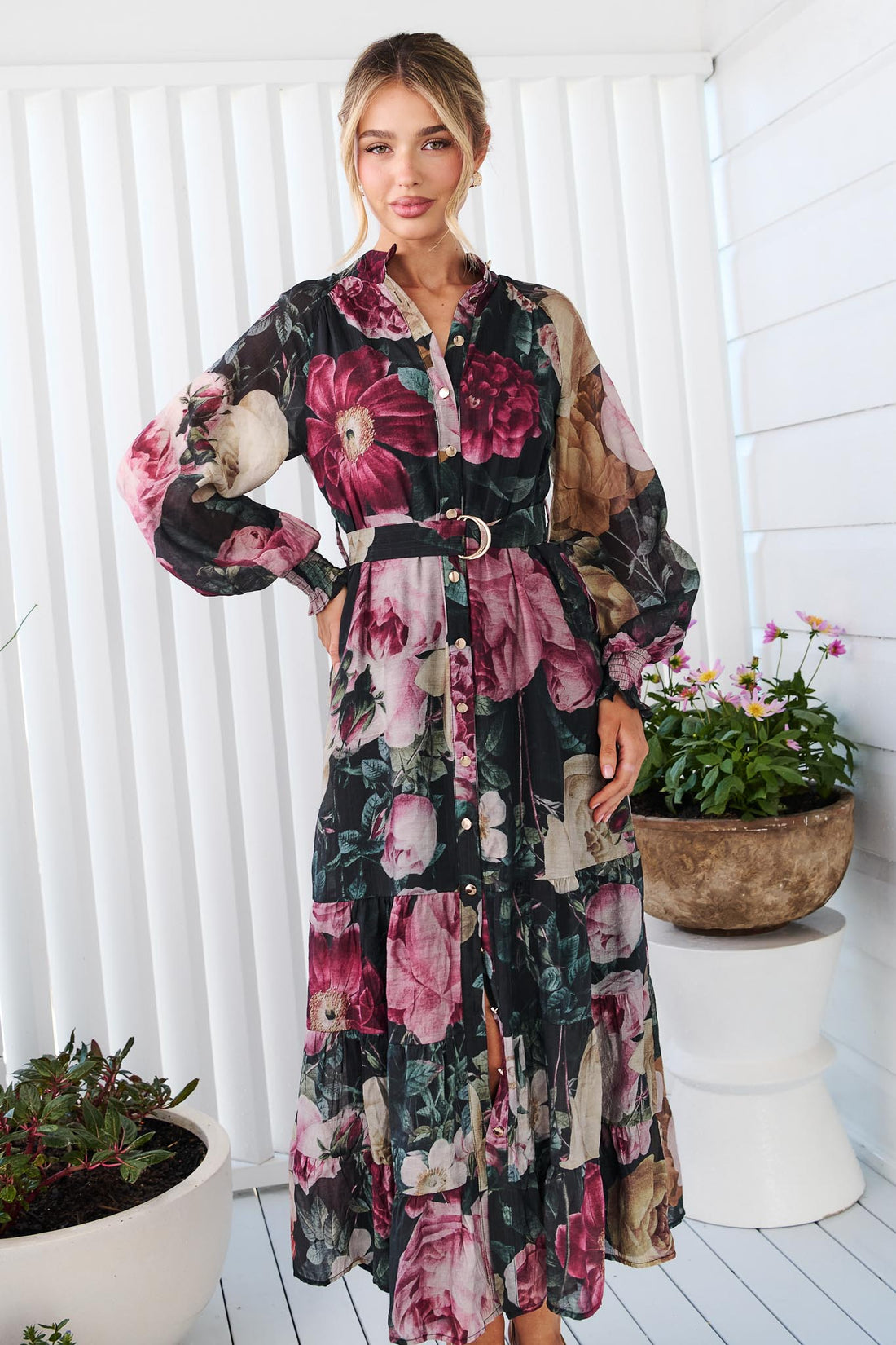 A woman wearing the minnie + MAXXIE Maison long-sleeved maxi event dress with a dark floral print featuring pink, red, and beige flowers on a black background, standing in front of a white panelled wall.