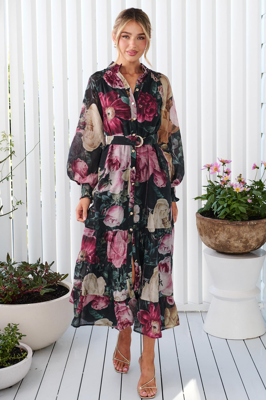 A woman wearing the minnie + MAXXIE Maison long-sleeved maxi event dress with a dark floral print featuring pink, red, and beige flowers on a black background, standing in front of a white panelled wall.