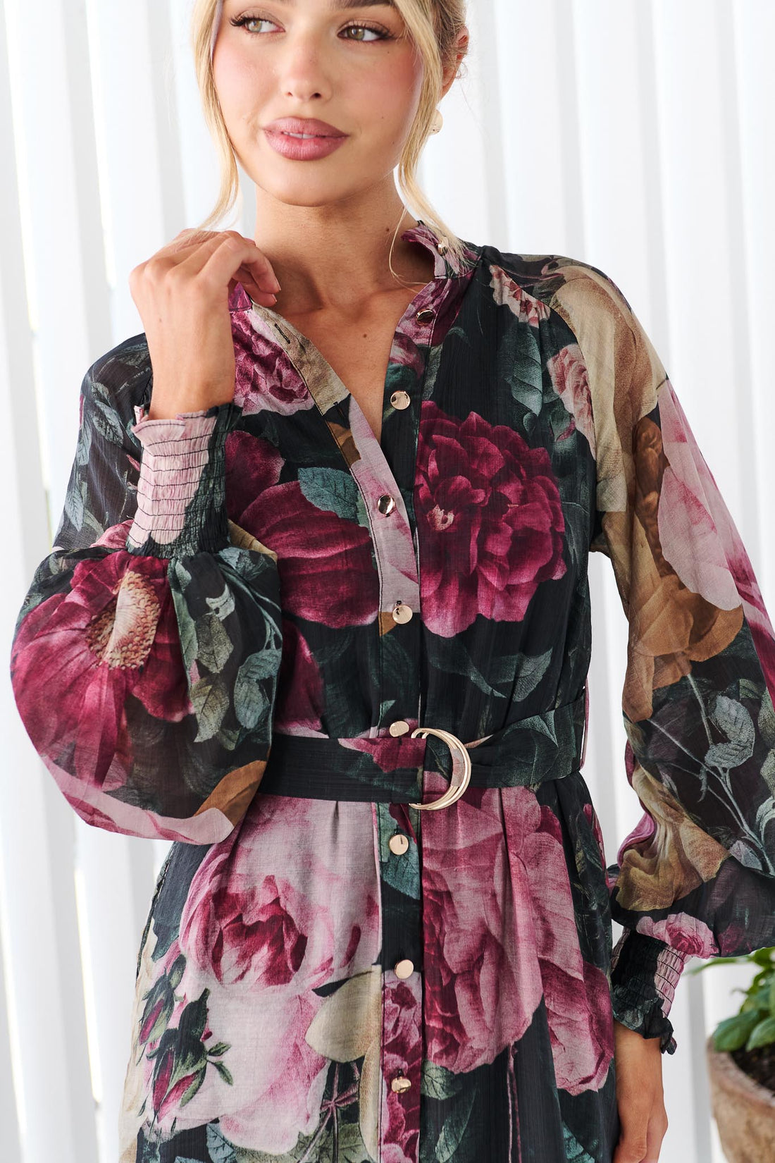Close up of a woman wearing the minnie + MAXXIE Maison long-sleeved maxi event dress with a dark floral print featuring pink, red, and beige flowers on a black background, standing in front of a white panelled wall.