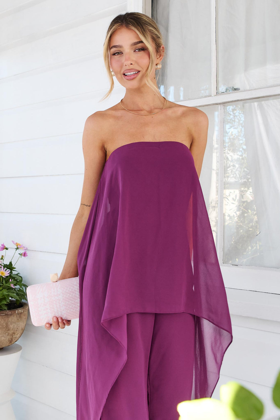 Close up of a woman wearing the minnie + MAXXIE Margot magenta jumpsuit, with sheer draped panels. She is standing on a white porch with a small pink clutch.