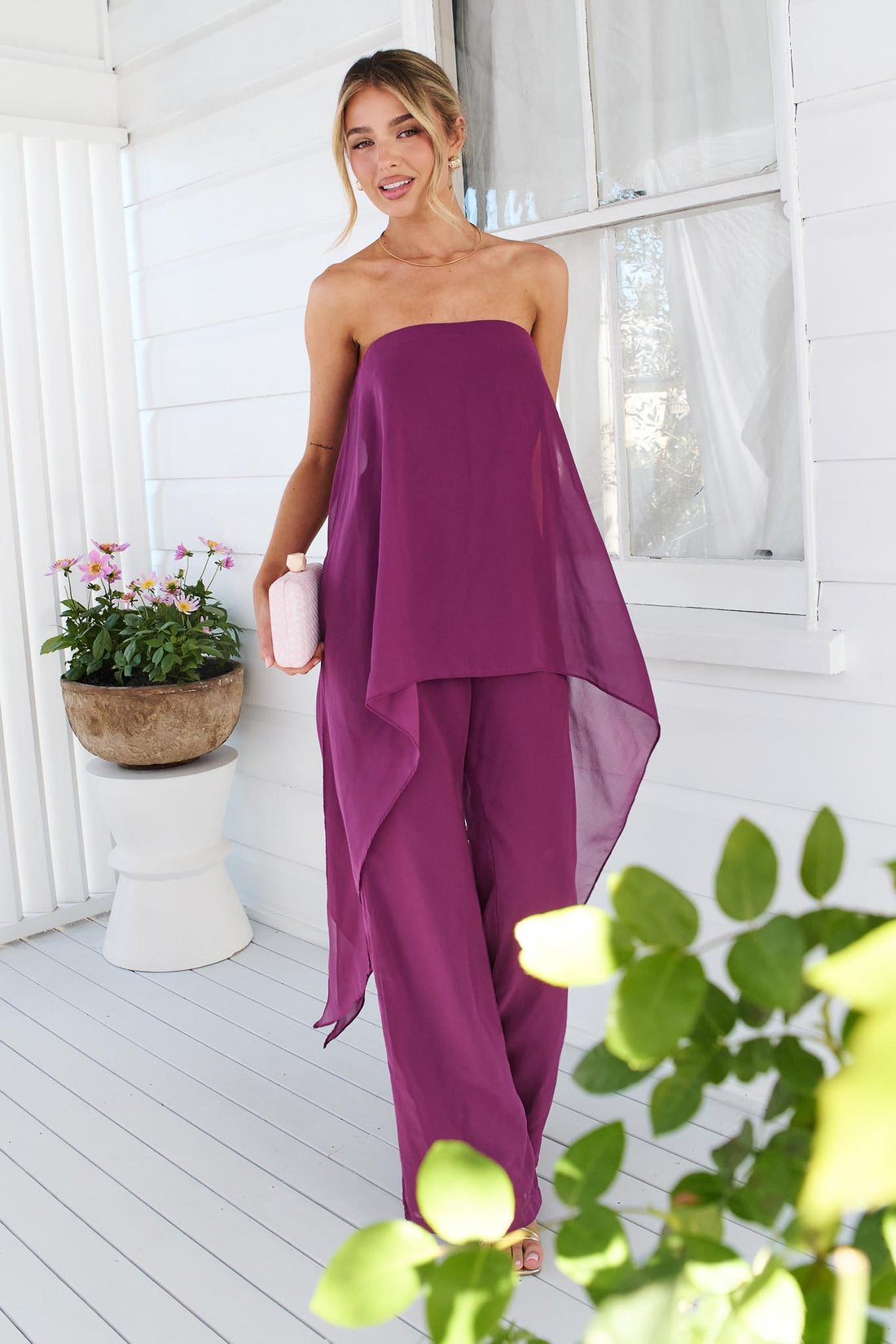 A woman wearing the minnie + MAXXIE Margot magenta jumpsuit, with sheer draped panels. She is standing on a white porch with a small pink clutch.