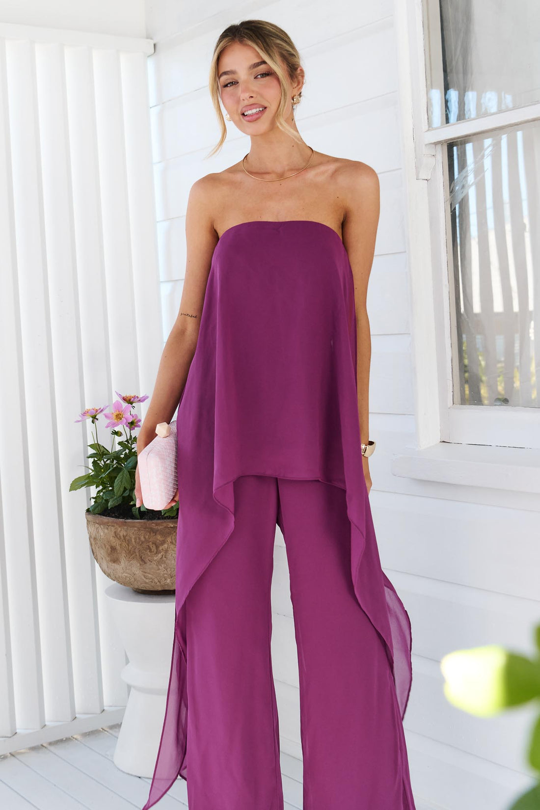 A woman wearing the minnie + MAXXIE Margot magenta jumpsuit, with sheer draped panels. She is standing on a white porch with a small pink clutch.
