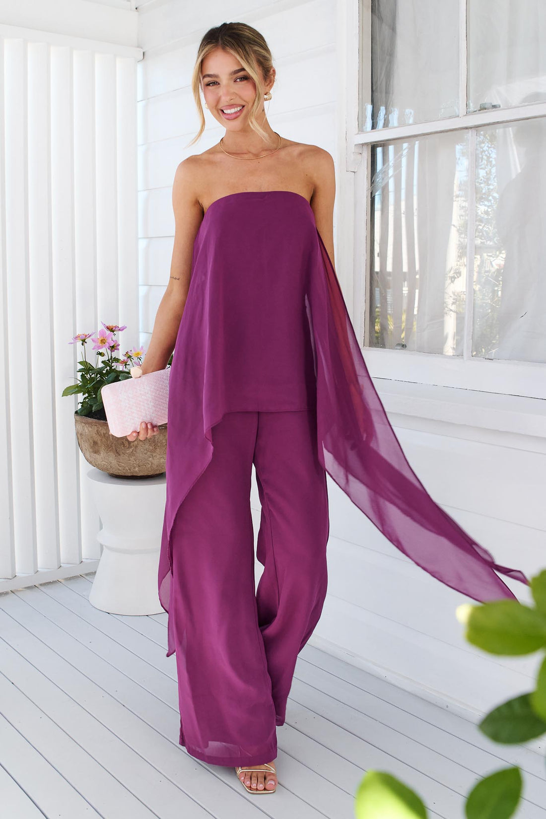 A woman wearing the minnie + MAXXIE Margot magenta jumpsuit, with sheer draped panels. She is standing on a white porch with a small pink clutch.