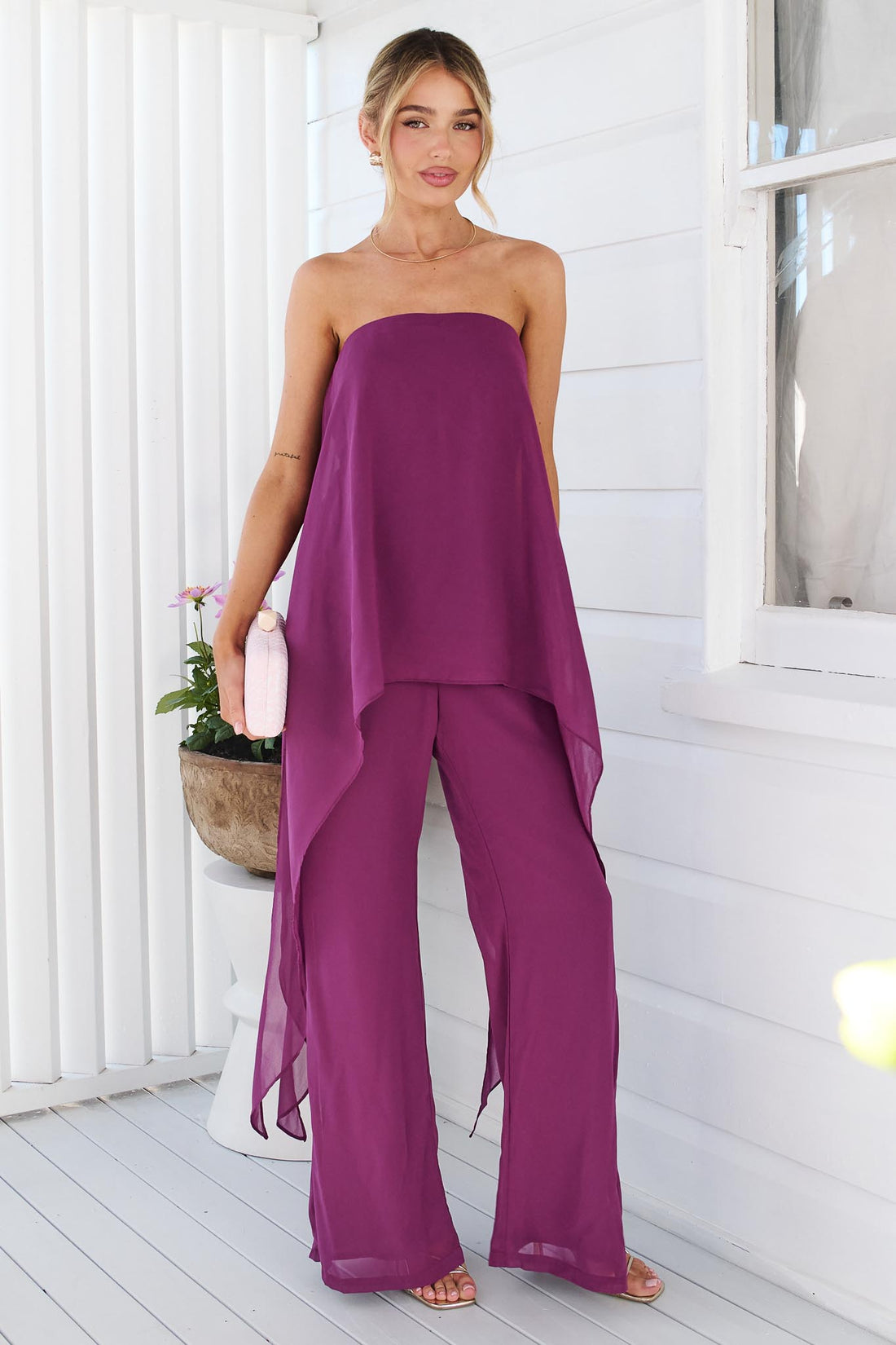 A woman wearing the minnie + MAXXIE Margot magenta jumpsuit, with sheer draped panels. She is standing on a white porch with a small pink clutch.