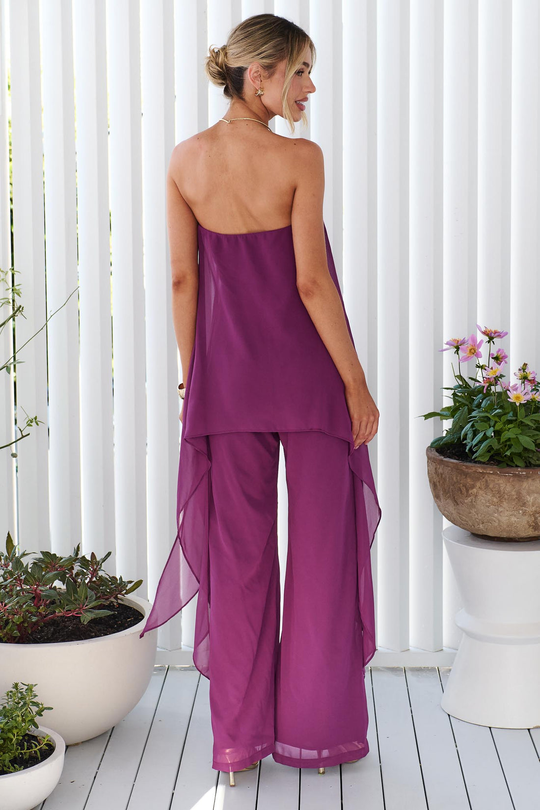 Back view of a woman wearing the minnie + MAXXIE Margot magenta jumpsuit, with sheer draped panels. She is standing on a white porch.