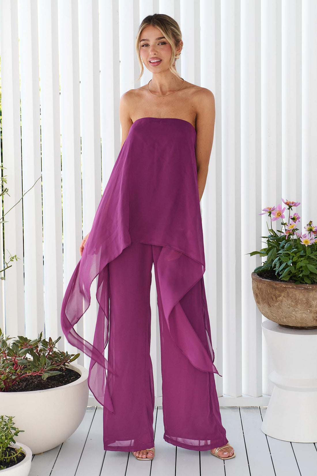 A woman wearing the minnie + MAXXIE Margot magenta jumpsuit, with sheer draped panels. She is standing on a white porch.