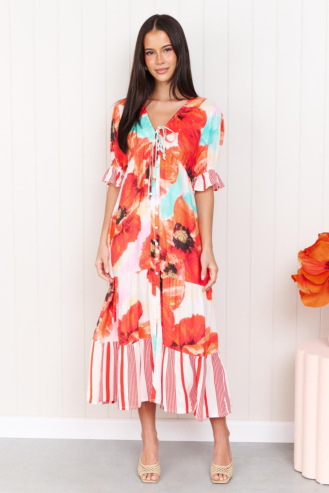 A woman wearing the minnie + MAXXIE Maryjane short-sleeve midi dress with a vibrant red, orange, and turquoise floral print on a white base, featuring ruffled sleeves and a tiered skirt, standing against a white panelled wall.