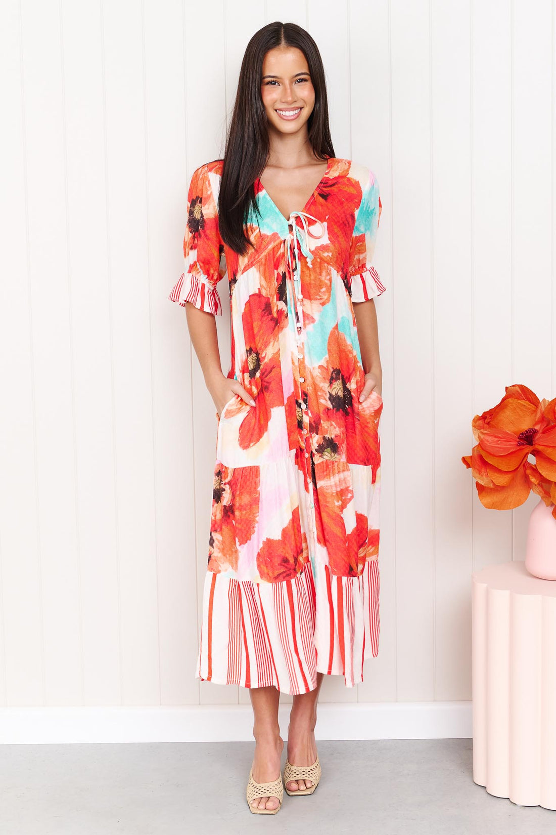 A woman wearing the minnie + MAXXIE Maryjane short-sleeve midi dress with a vibrant red, orange, and turquoise floral print on a white base, featuring ruffled sleeves and a tiered skirt, standing against a white panelled wall.