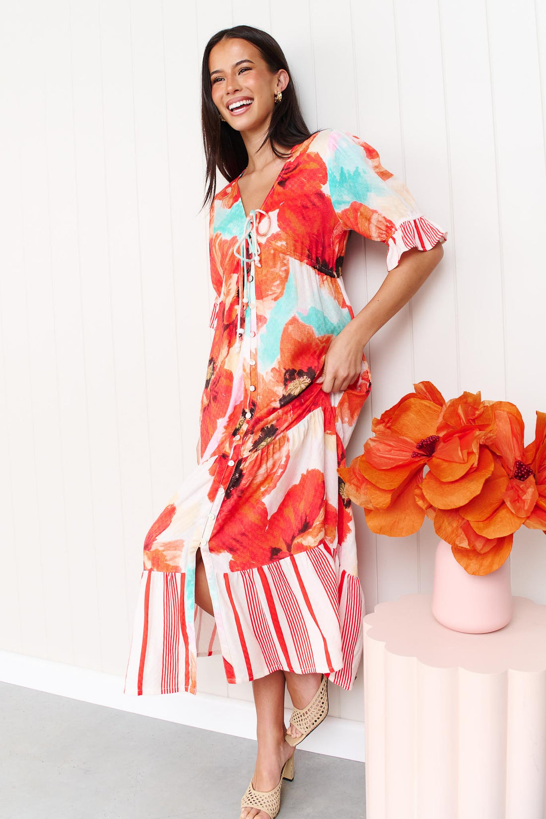 A woman wearing the minnie + MAXXIE Maryjane short-sleeve midi dress with a vibrant red, orange, and turquoise floral print on a white base, featuring ruffled sleeves and a tiered skirt, standing against a white panelled wall.