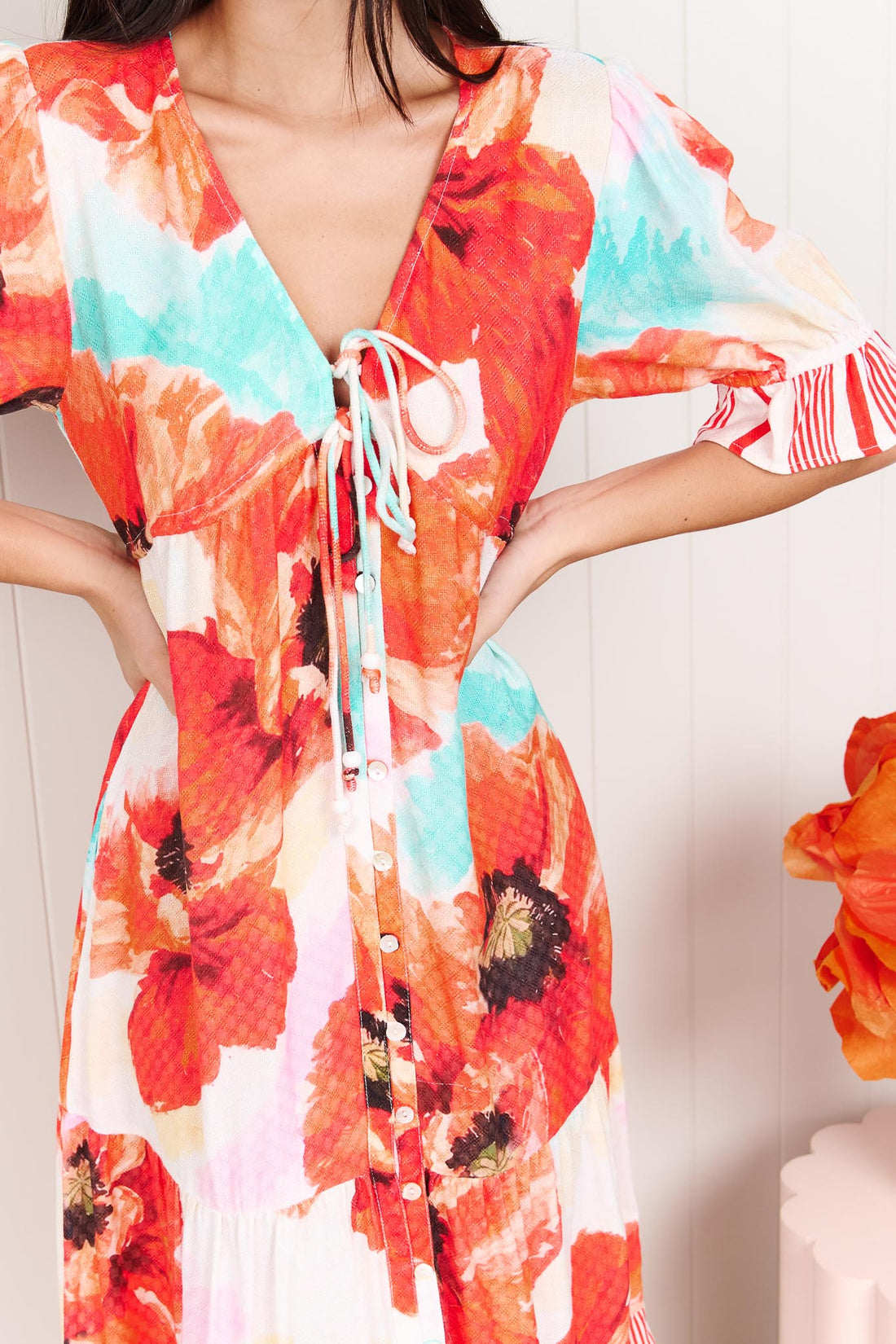 Close up of a woman wearing the minnie + MAXXIE Maryjane short-sleeve midi dress with a vibrant red, orange, and turquoise floral print on a white base, featuring ruffled sleeves and a tiered skirt, standing against a white panelled wall.
