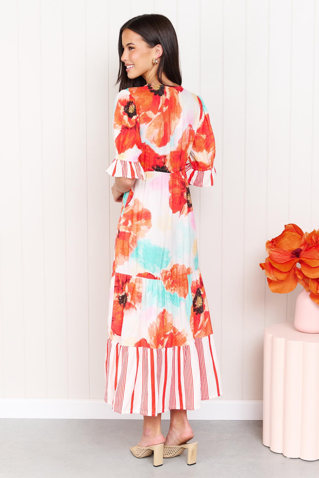 Back view of a woman wearing the minnie + MAXXIE Maryjane short-sleeve midi dress with a vibrant red, orange, and turquoise floral print on a white base, featuring ruffled sleeves and a tiered skirt, standing against a white panelled wall.