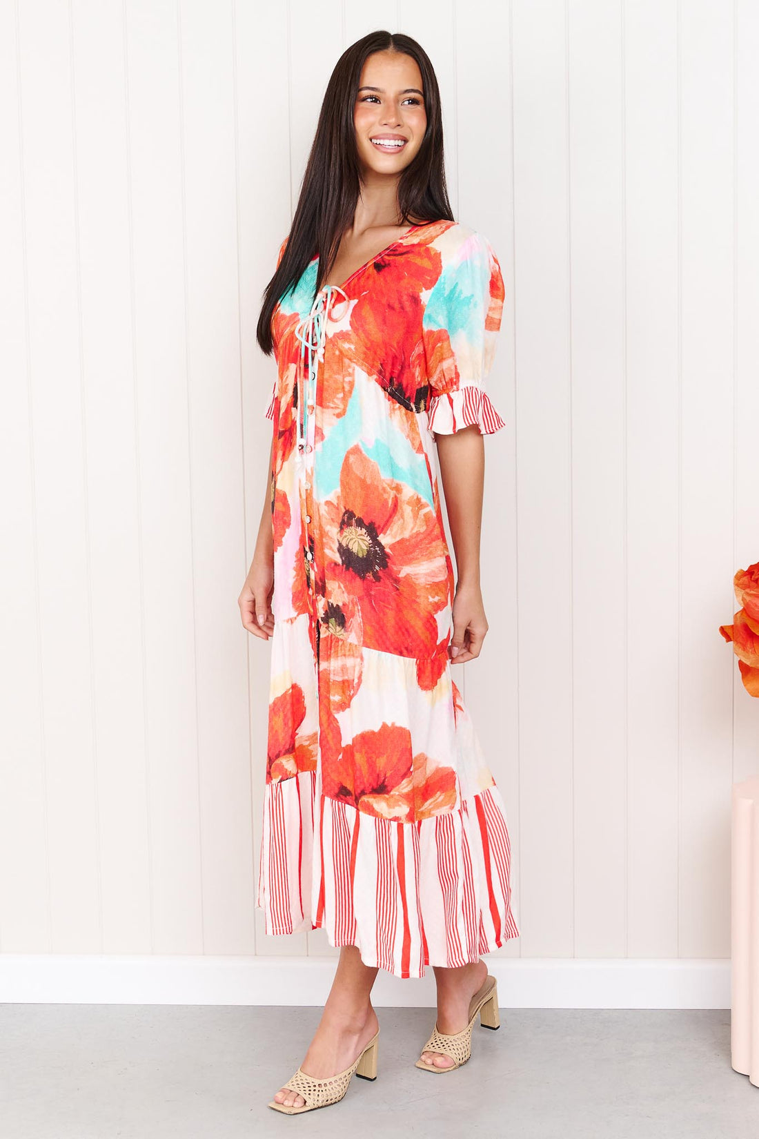 A woman wearing the minnie + MAXXIE Maryjane short-sleeve midi dress with a vibrant red, orange, and turquoise floral print on a white base, featuring ruffled sleeves and a tiered skirt, standing against a white panelled wall.
