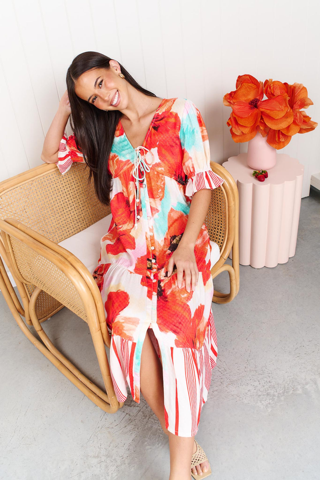A woman wearing the minnie + MAXXIE Maryjane short-sleeve midi dress with a vibrant red, orange, and turquoise floral print on a white base, featuring ruffled sleeves and a tiered skirt, sitting on a rattan chair.