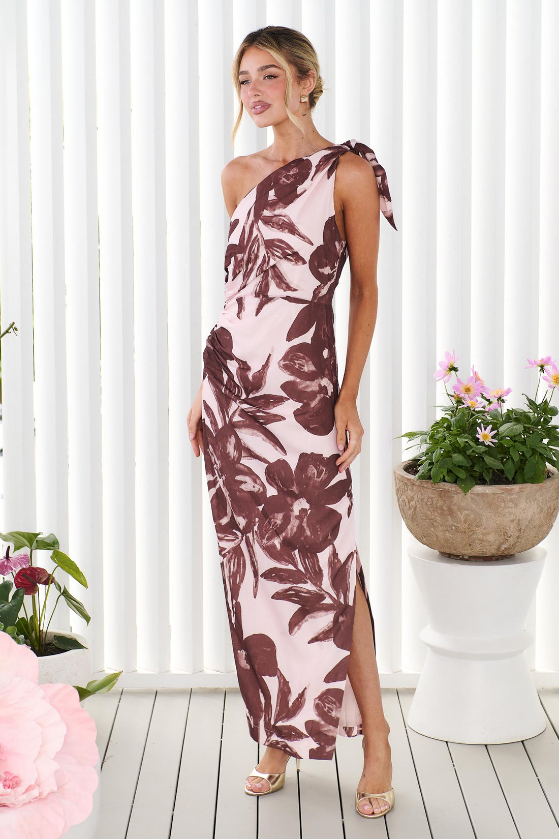 A woman wearing the minnie + MAXXIE Maya a one-shoulder maxi dress with a brown floral print on a light pink background, set against a bright white slatted wall with potted plants.