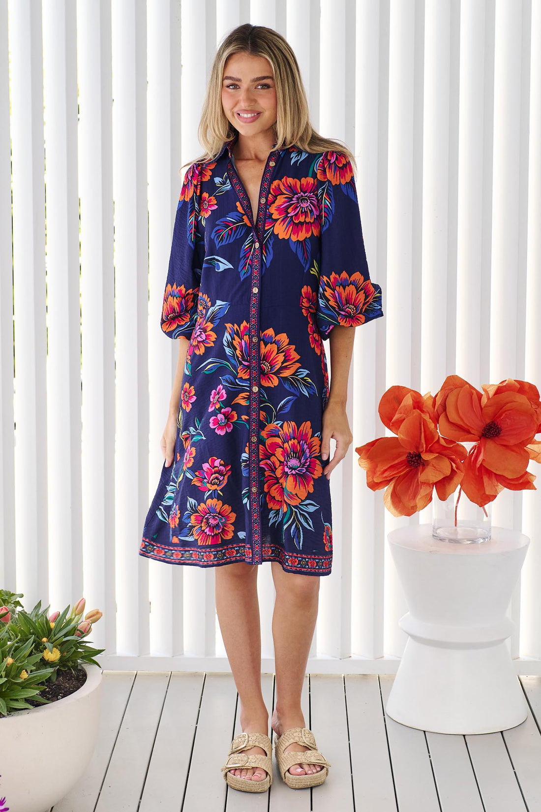 A woman wearing the minnie + MAXXIE Mia Dress in navy floral with vibrant orange and pink flowers, set against a white slatted background.