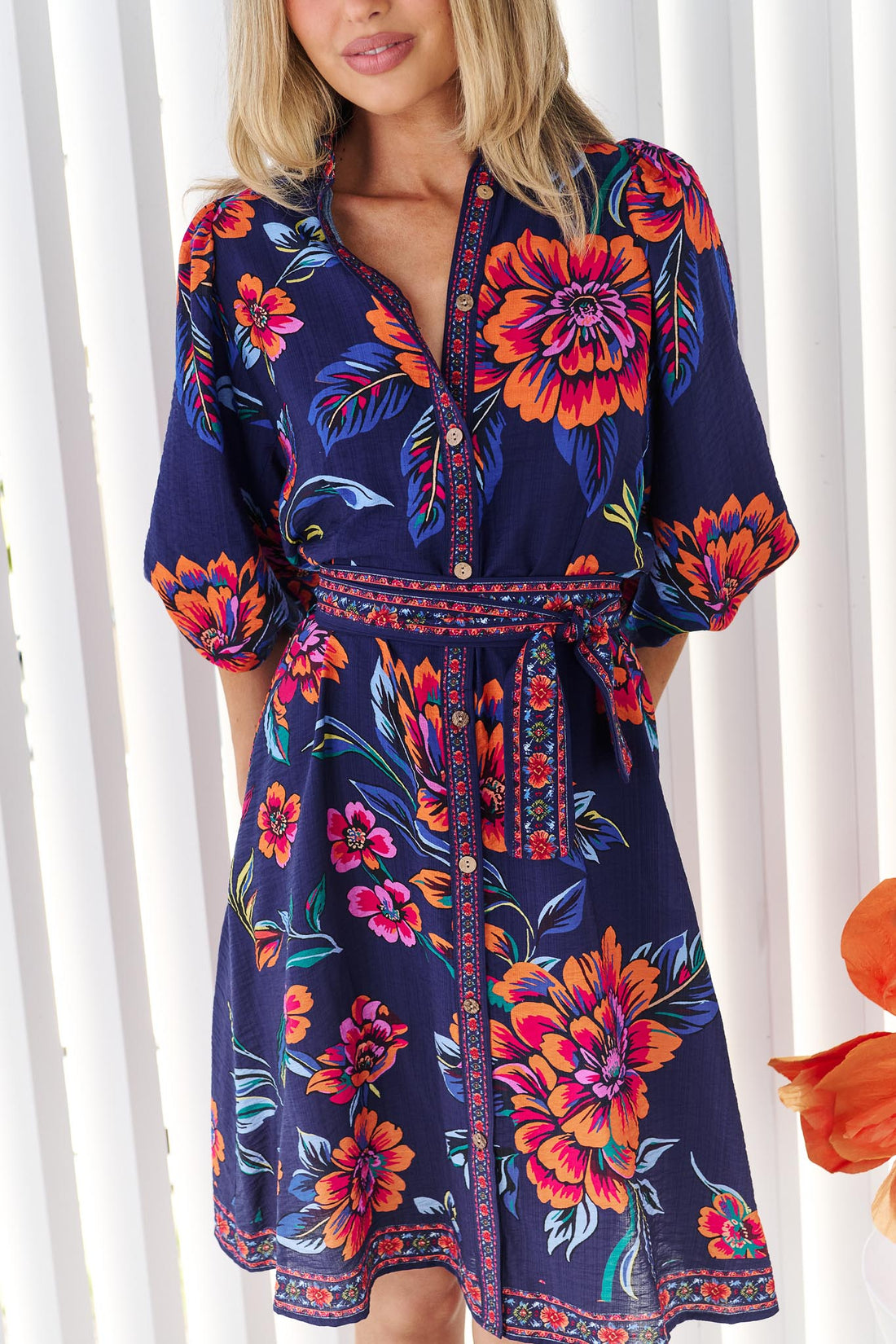 Close up of a woman wearing the minnie + MAXXIE Mia Dress in navy floral with vibrant orange and pink flowers and matching waist tie, set against a white slatted background.
