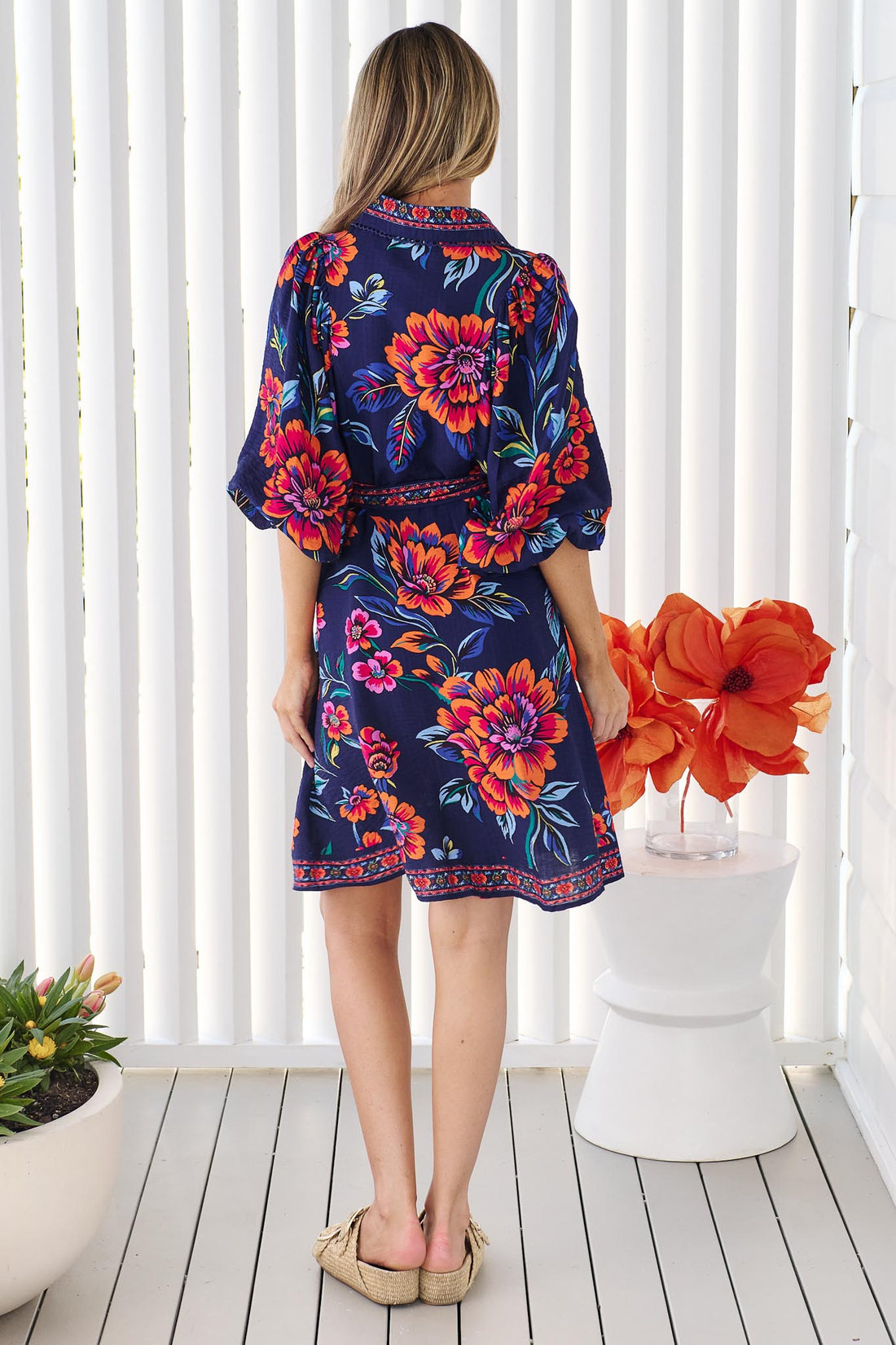 Back view of a woman wearing the minnie + MAXXIE Mia Dress in navy floral with vibrant orange and pink flowers and matching waist tie, set against a white slatted background.