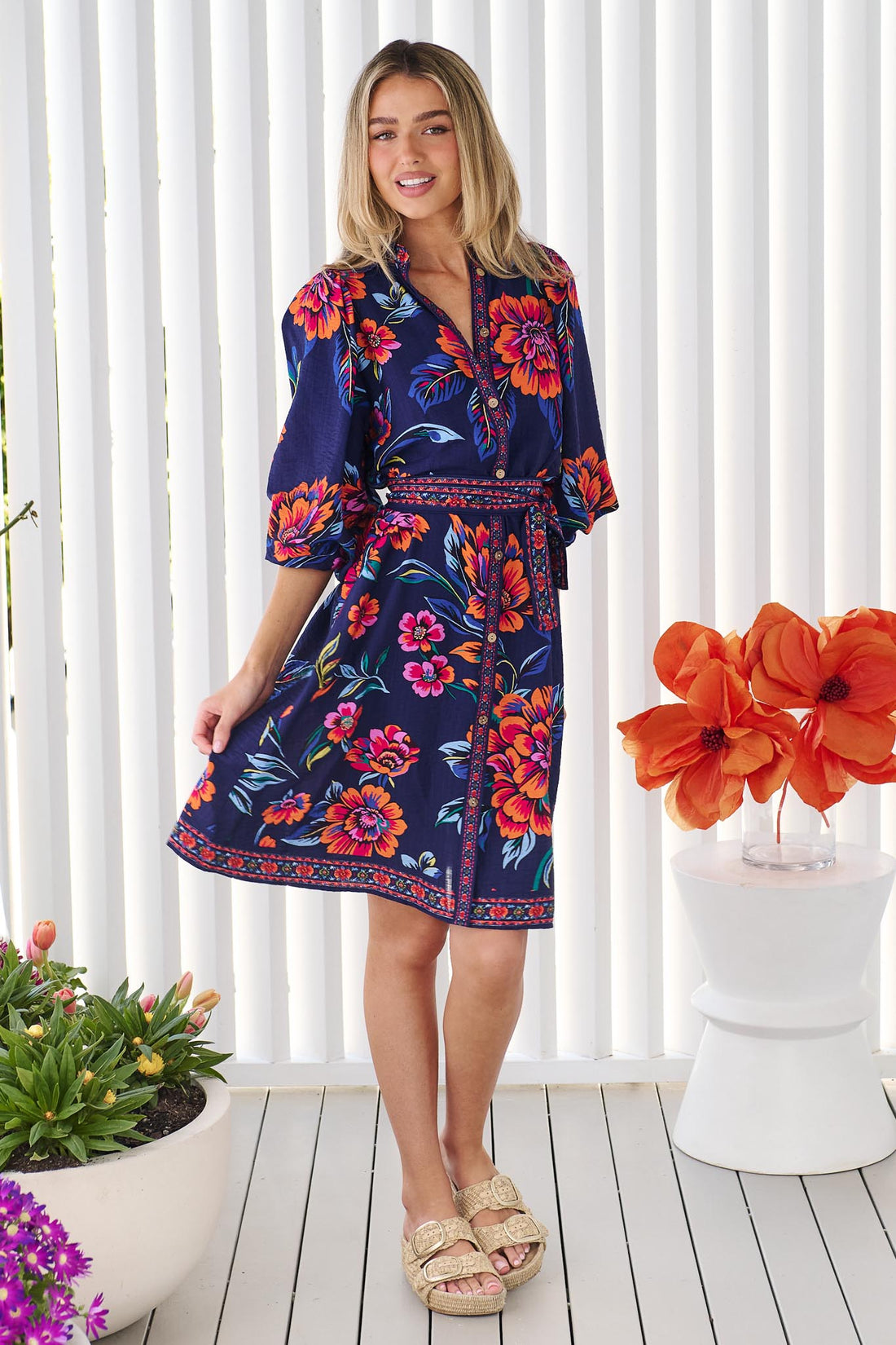 A woman wearing the minnie + MAXXIE Mia Dress in navy floral with vibrant orange and pink flowers and matching waist tie, set against a white slatted background.