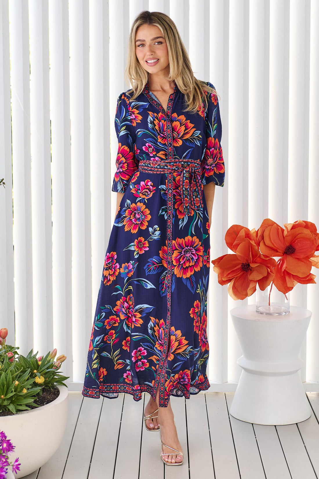 A woman wearing the minnie + MAXXIE Mia maxi shirt dress in navy blue with a vibrant pink, orange, and blue floral print, and matching waist tie, standing in front of a white panelled wall.