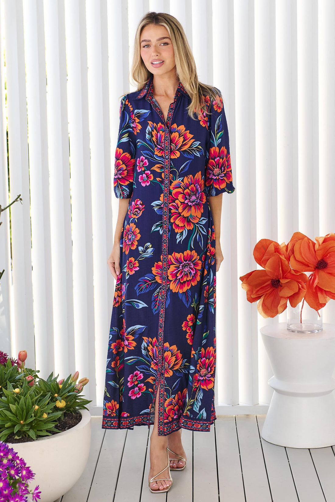 A woman wearing the minnie + MAXXIE Mia maxi shirt dress in navy blue with a vibrant pink, orange, and blue floral print, standing in front of a white panelled wall.