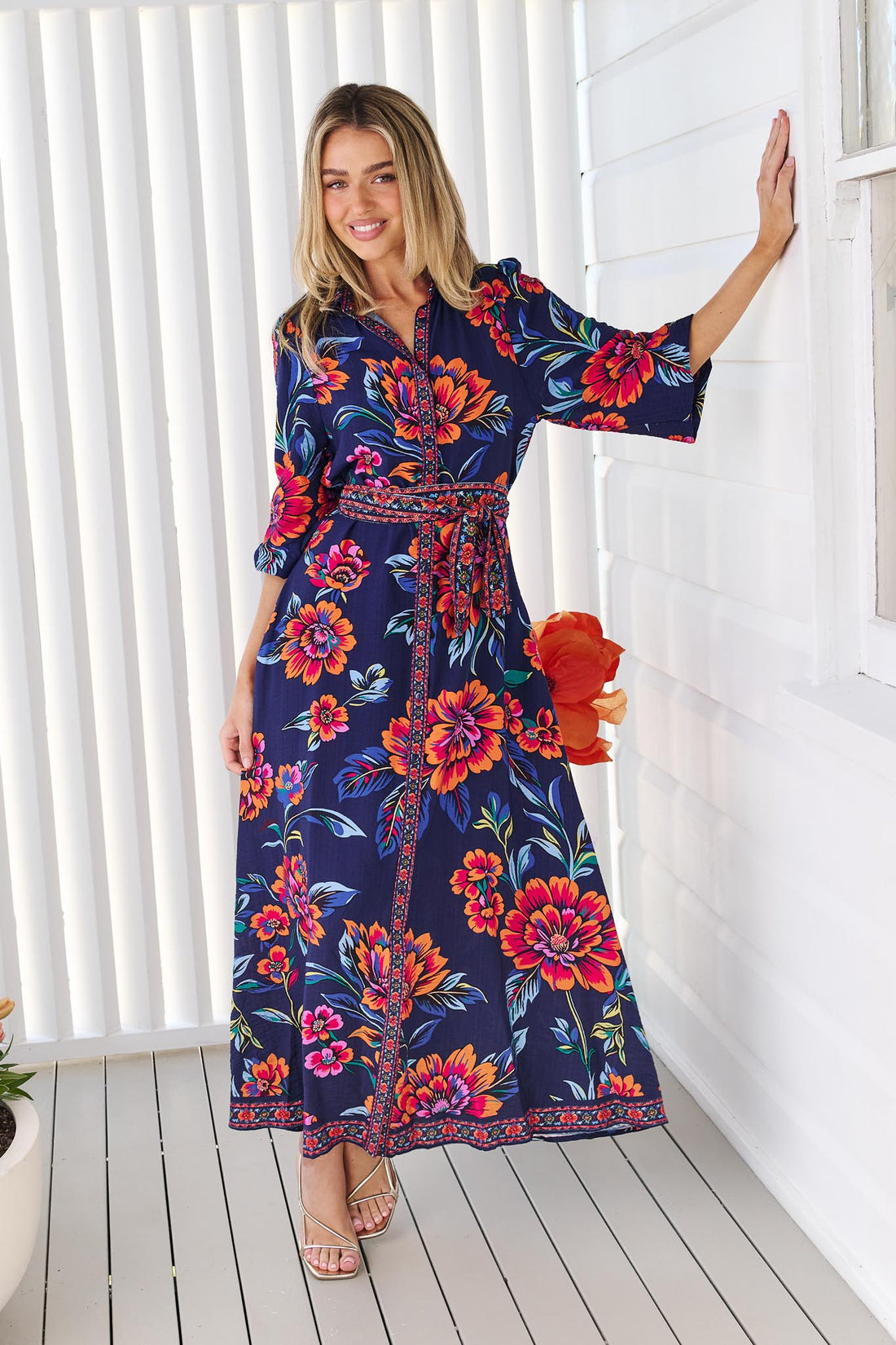 A woman wearing the minnie + MAXXIE Mia maxi shirt dress in navy blue with a vibrant pink, orange, and blue floral print, and matching waist tie, standing in front of a white panelled wall.