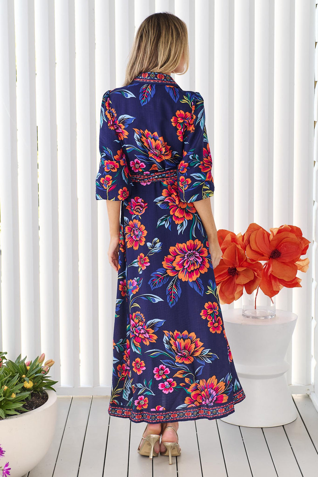 Back view of a woman wearing the minnie + MAXXIE Mia maxi shirt dress in navy blue with a vibrant pink, orange, and blue floral print, and matching waist tie, standing in front of a white panelled wall.