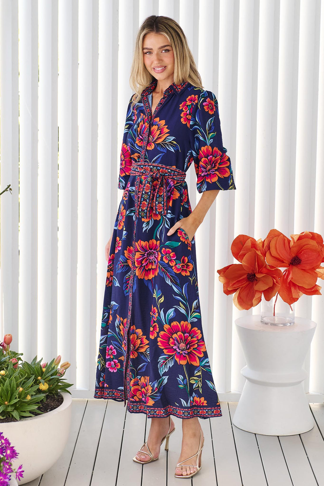 Side view of a woman wearing the minnie + MAXXIE Mia maxi shirt dress in navy blue with a vibrant pink, orange, and blue floral print, and matching waist tie, standing in front of a white panelled wall.