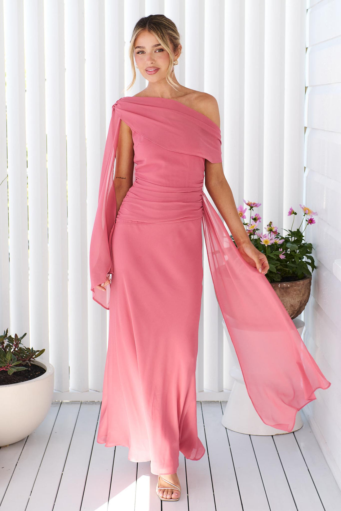 A woman wearing the minnie + MAXXIE Miro rose pink one-shoulder maxi dress with a flowing chiffon sash, standing in front of a white panelled wall.