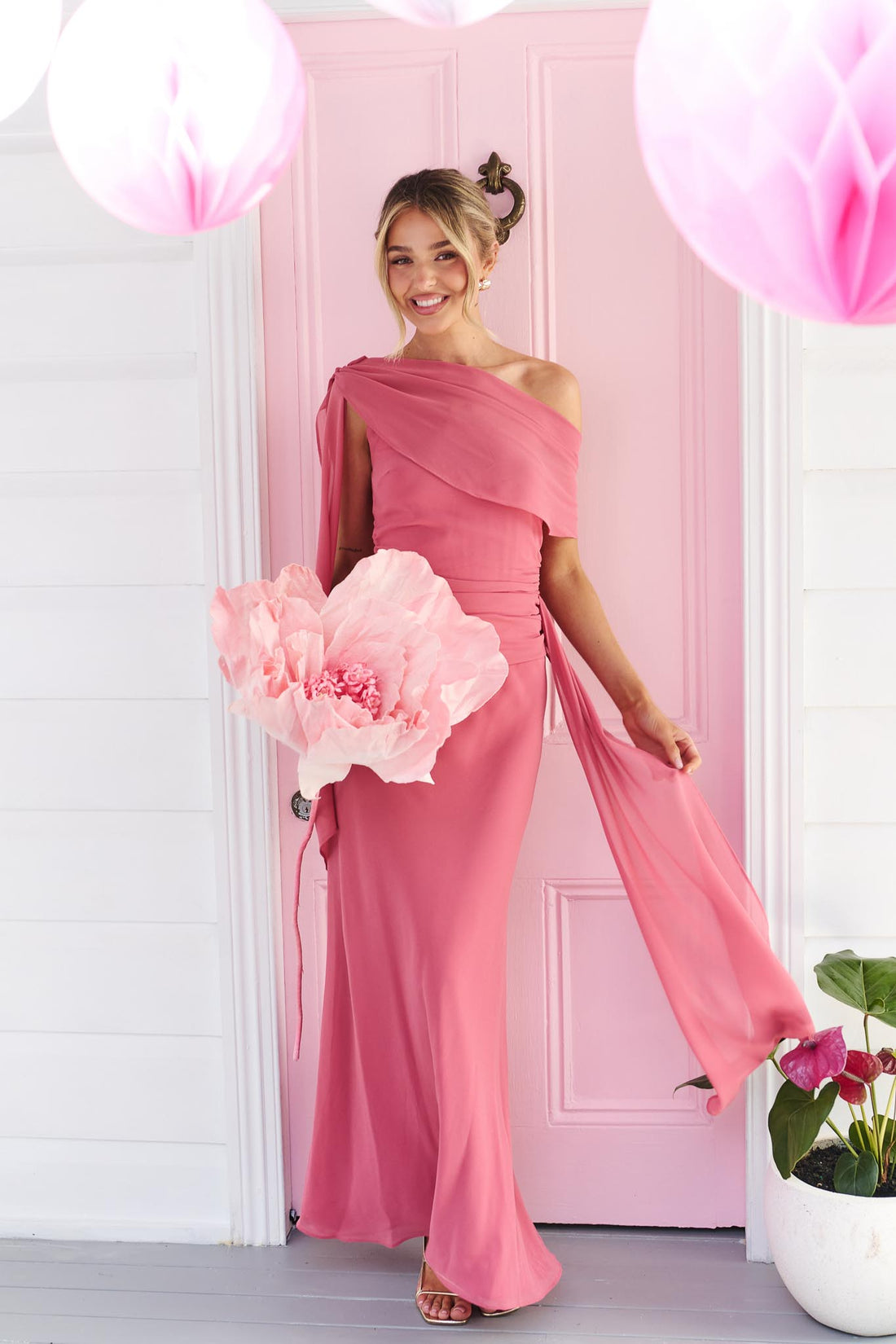 A woman wearing the minnie + MAXXIE Miro rose pink one-shoulder maxi dress with a flowing chiffon sash, standing in front of a pink door.