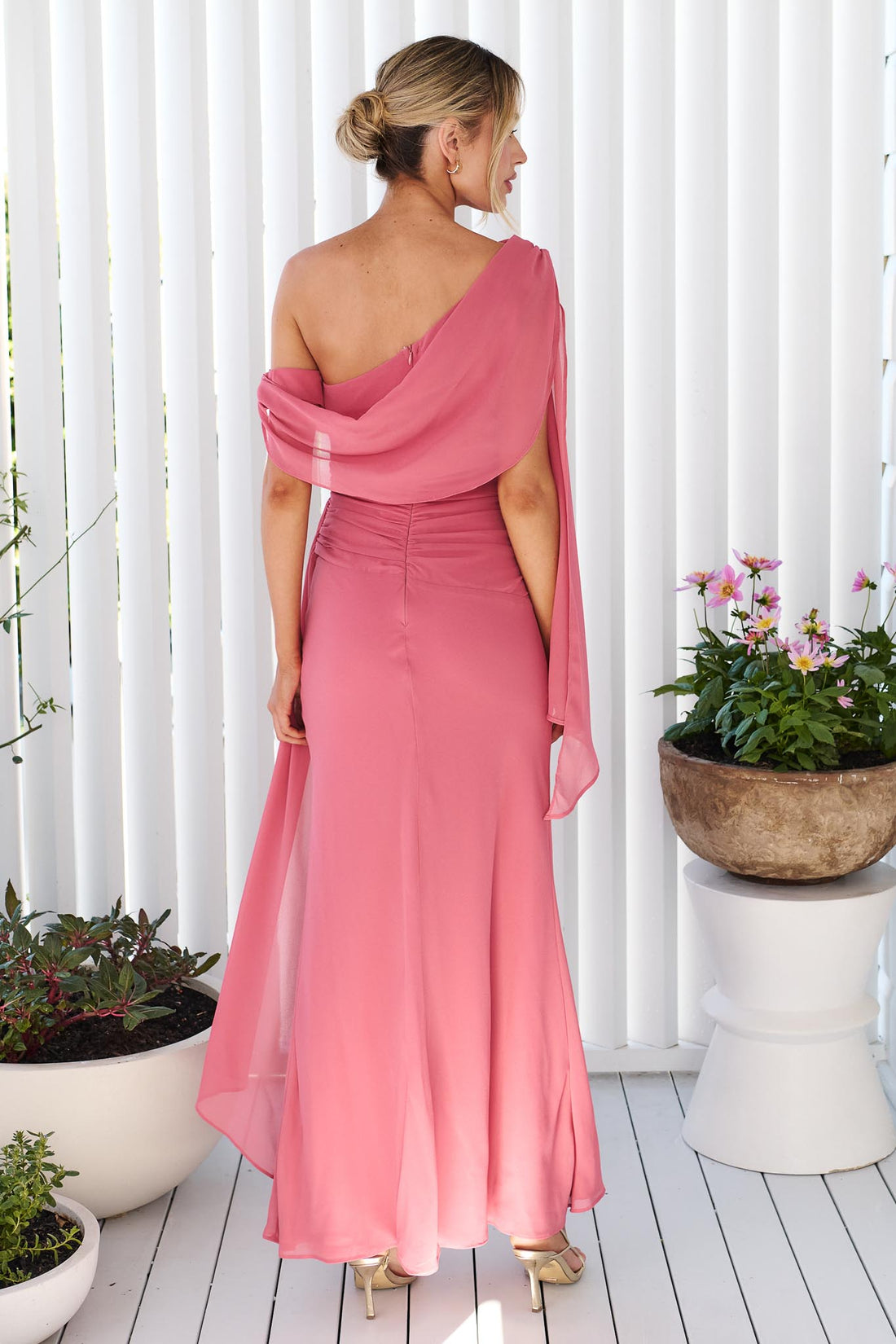 Back view of a woman wearing the minnie + MAXXIE Miro rose pink one-shoulder maxi dress with a flowing chiffon sash, standing in front of a white panelled wall.