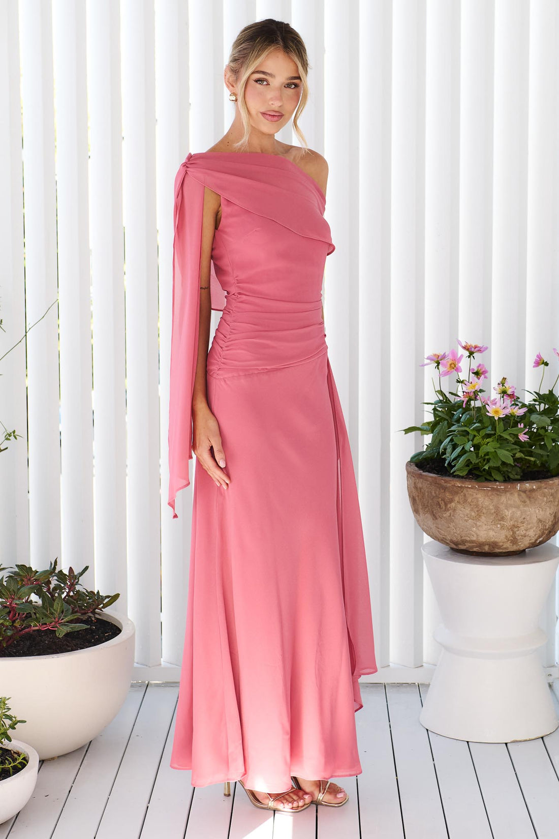 A woman wearing the minnie + MAXXIE Miro rose pink one-shoulder maxi dress with a flowing chiffon sash, standing in front of a white panelled wall.