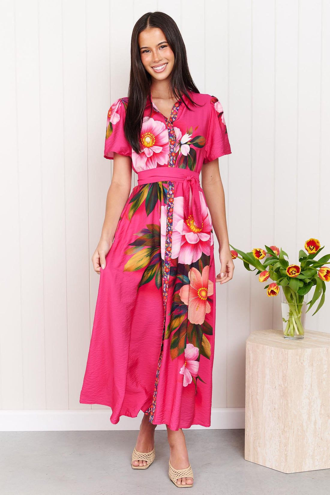 A woman wearing the minnie + MAXXIE Mona Maxi Dress in bright fuchsia pink with a large floral print and waist tie, standing in front of a white panelled wall.