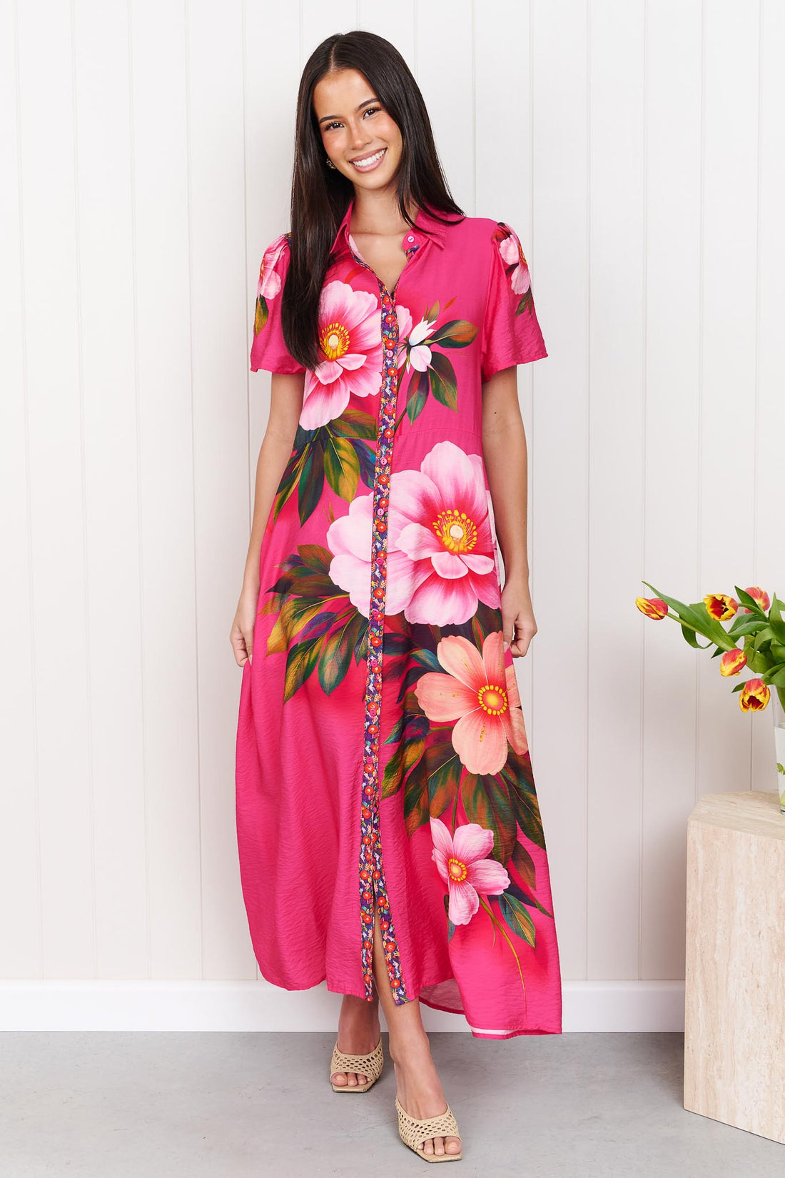 A woman wearing the minnie + MAXXIE Mona Maxi Dress in bright fuchsia pink with a large floral print, standing in front of a white panelled wall.