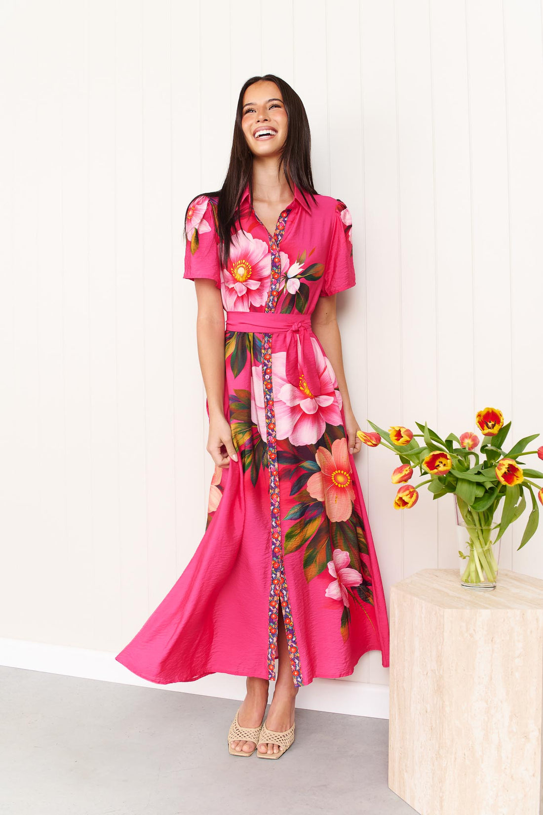 A woman wearing the minnie + MAXXIE Mona Maxi Dress in bright fuchsia pink with a large floral print and waist tie, standing in front of a white panelled wall with a vase of orange and yellow tulips.