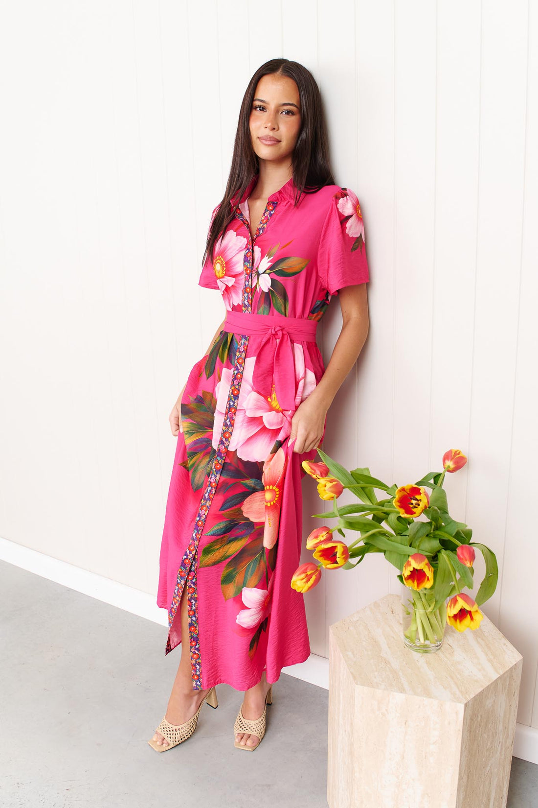 A woman wearing the minnie + MAXXIE Mona Maxi Dress in bright fuchsia pink with a large floral print and waist tie, standing in front of a white panelled wall with a vase of orange and yellow tulips.