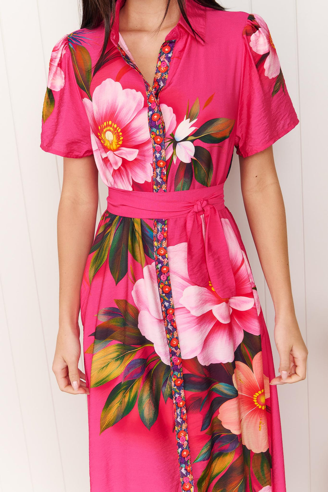 Detail of a woman wearing the minnie + MAXXIE Mona Maxi Dress in bright fuchsia pink with a large floral print and waist tie, standing in front of a white panelled wall.