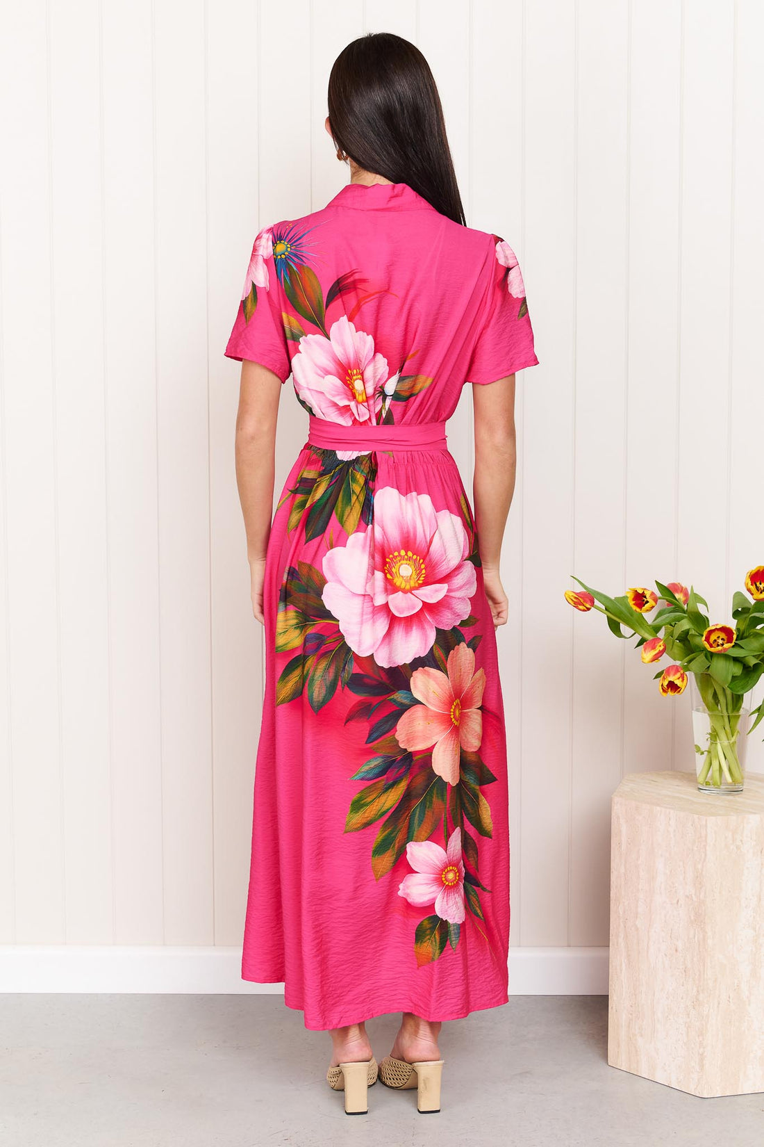 Back view of a woman wearing the minnie + MAXXIE Mona Maxi Dress in bright fuchsia pink with a large floral print and waist tie, standing in front of a white panelled wall.