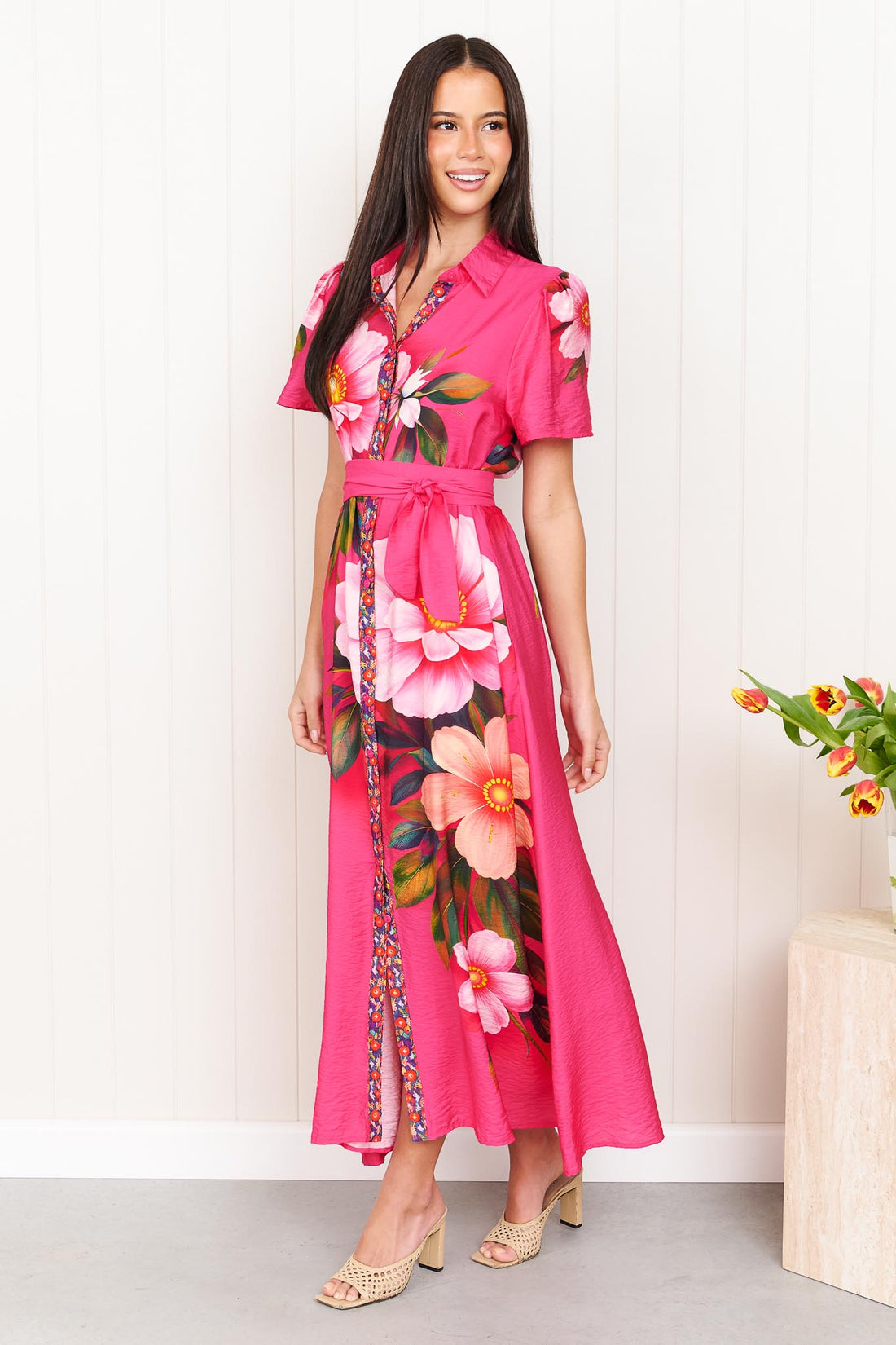 A woman wearing the minnie + MAXXIE Mona Maxi Dress in bright fuchsia pink with a large floral print and waist tie, standing in front of a white panelled wall.