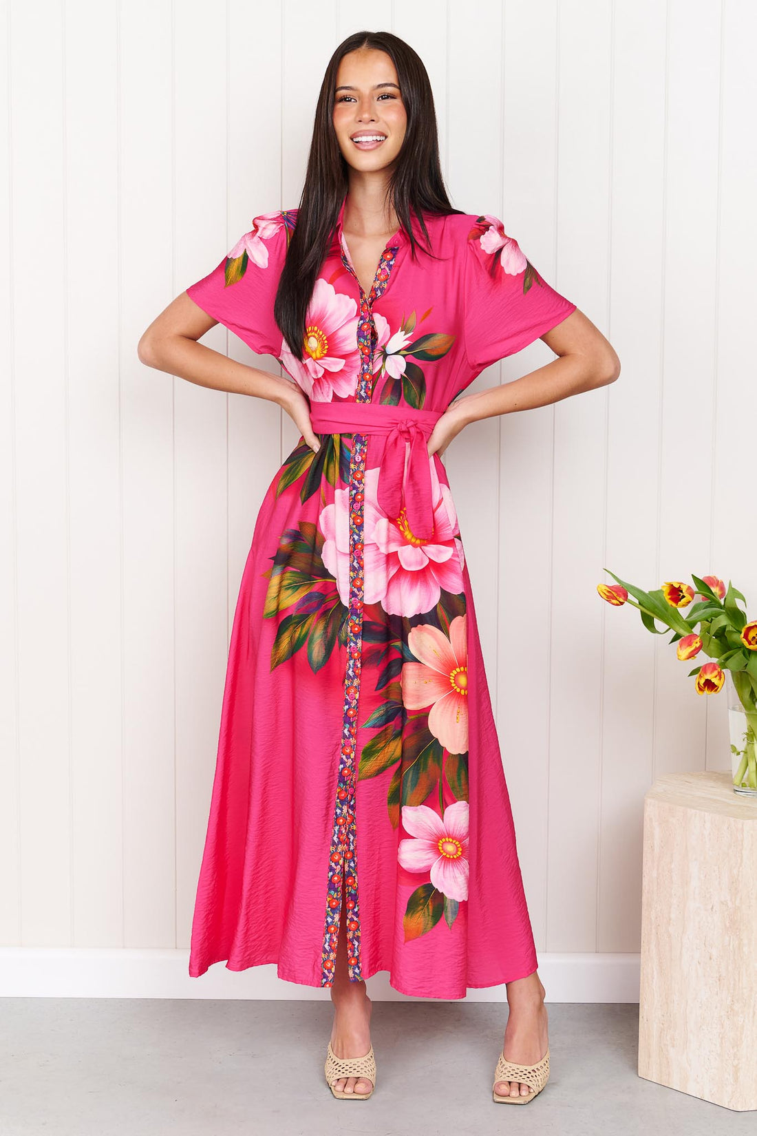 A woman wearing the minnie + MAXXIE Mona Maxi Dress in bright fuchsia pink with a large floral print and waist tie, standing in front of a white panelled wall.