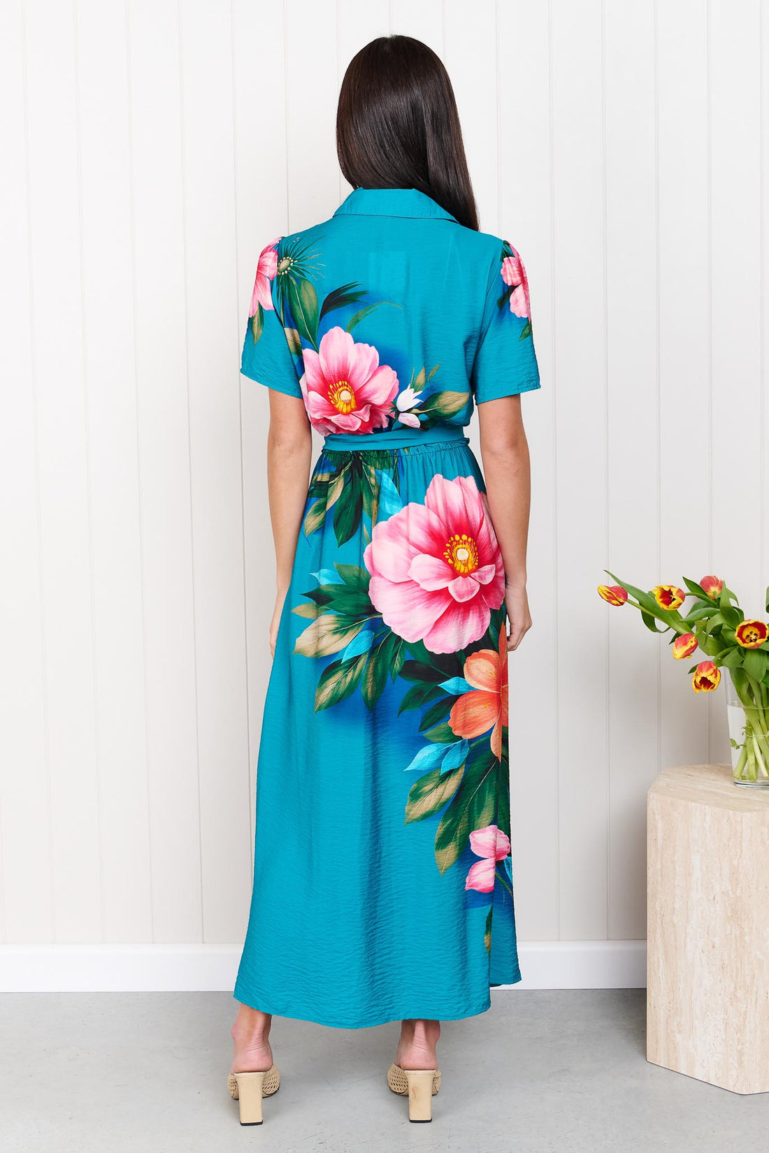 Back view of a woman wearing the minnie + MAXXIE Mona Maxi Dress in bright teal blue with a large floral print and waist tie, standing in front of a white panelled wall.